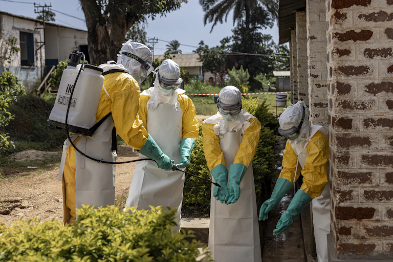 Four health workers stand in front of a brick building in head-to-toe protective gear as one disinfects the others' gloves.