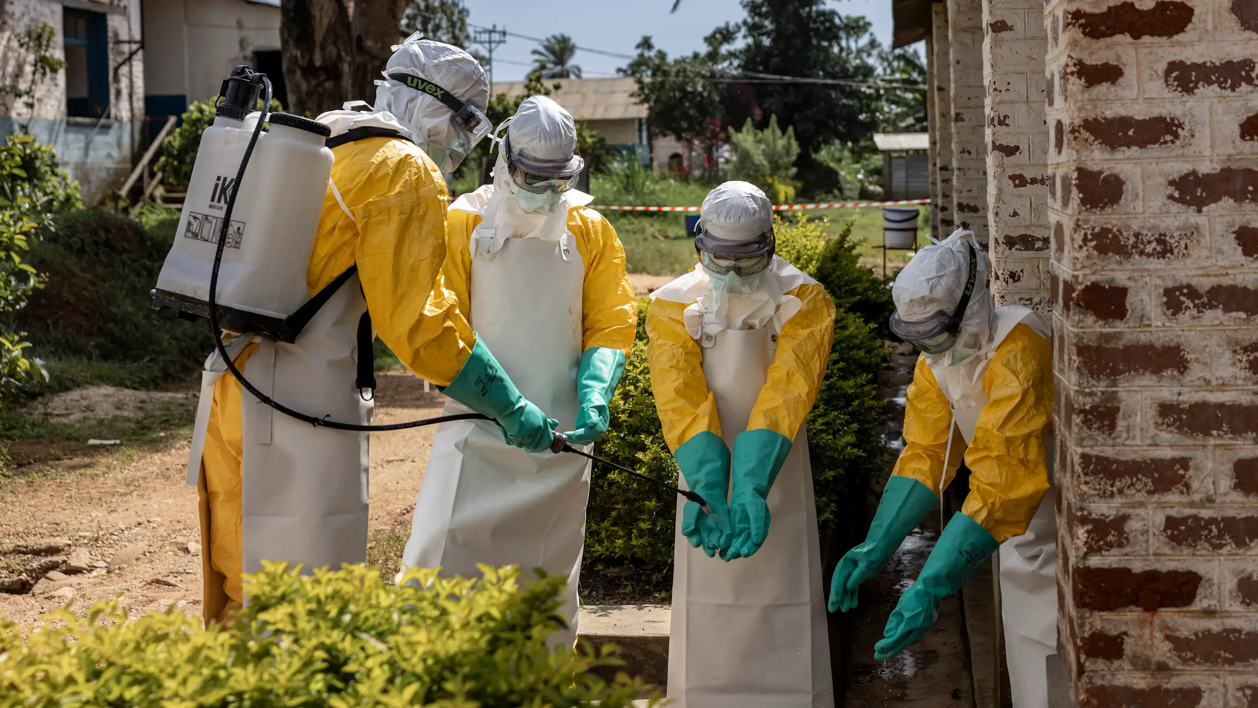 Four health workers stand in front of a brick building in head-to-toe protective gear as one disinfects the others' gloves.