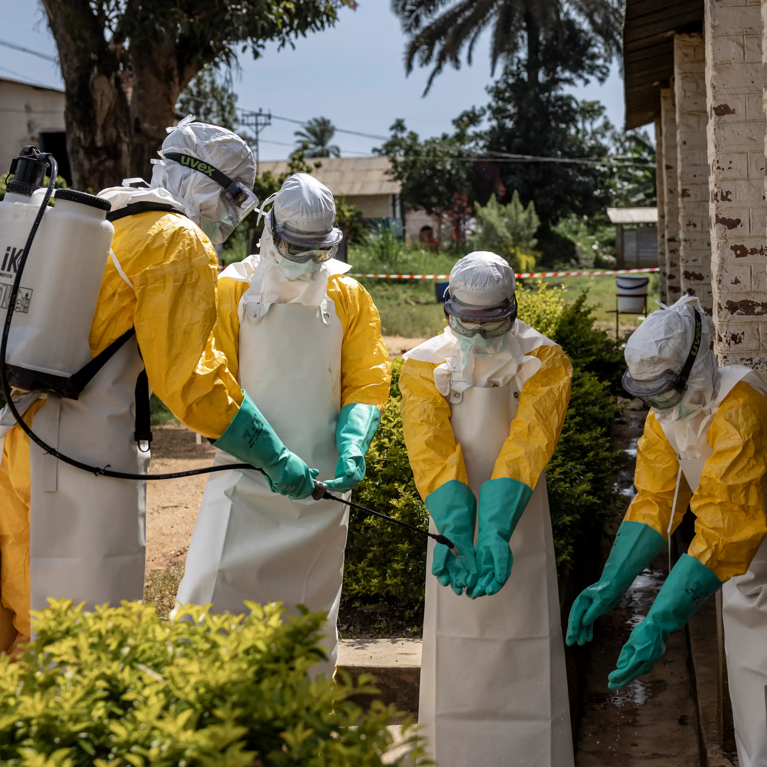 Four health workers stand in front of a brick building in head-to-toe protective gear as one disinfects the others' gloves.
