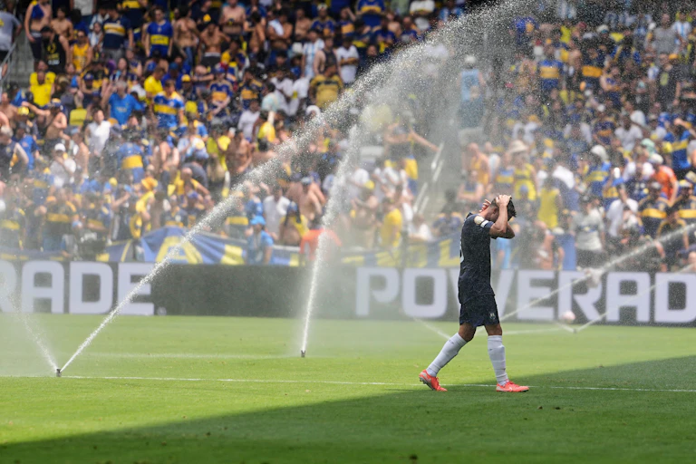 A player in a black uniform and orange shoes walks through sprinklers on a soccer field