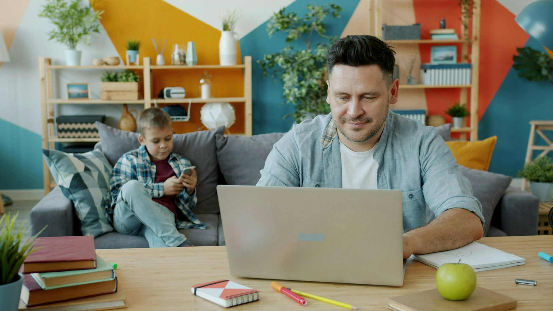 Un hombre de mediana edad trabaja en una computadora portátil mientras un niño se sienta en un sofá al fondo.