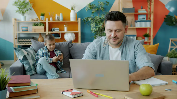 El matrimonio, el divorcio y la paternidad influyen en las decisiones de los canadienses de trabajar por cuenta propia: así es como 1 Un hombre de mediana edad trabaja en una computadora portátil mientras un niño se sienta en un sofá al fondo.