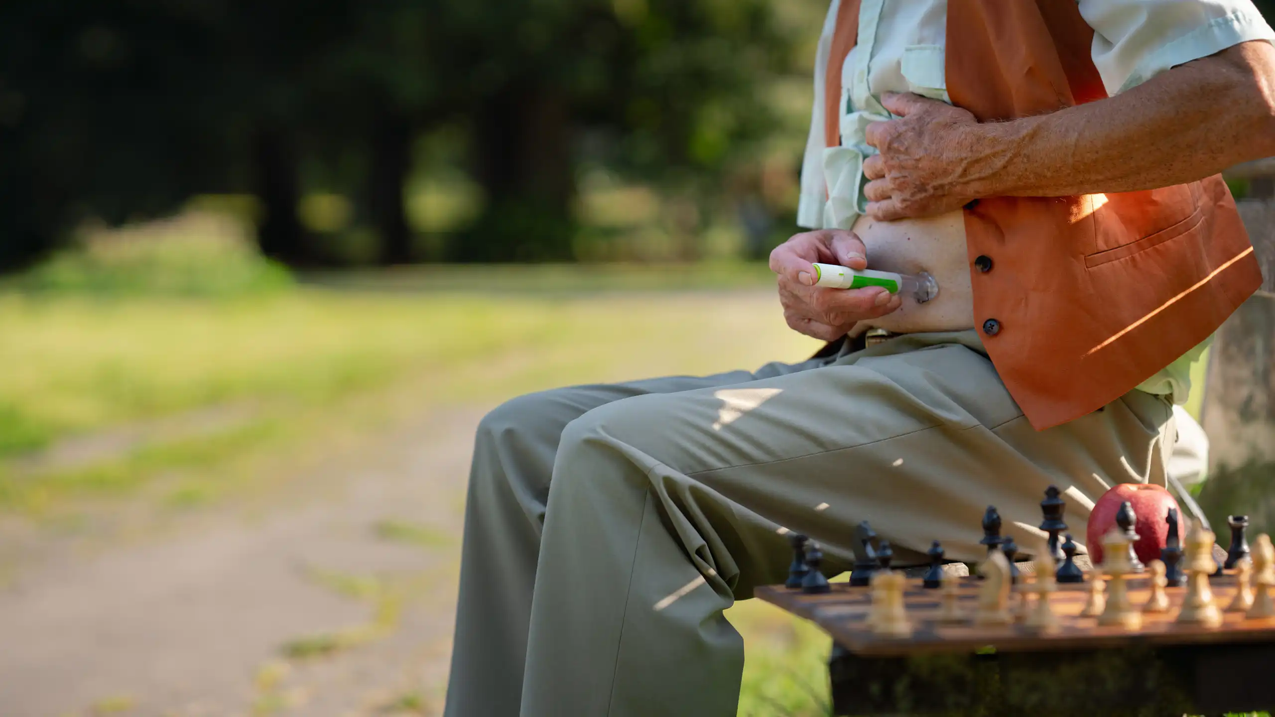An elderly man sat on a park bench injects himself with insulin.