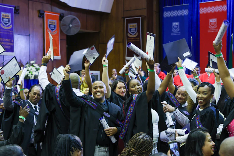 Smiling young people in academic gowns, raising certificates above their heads