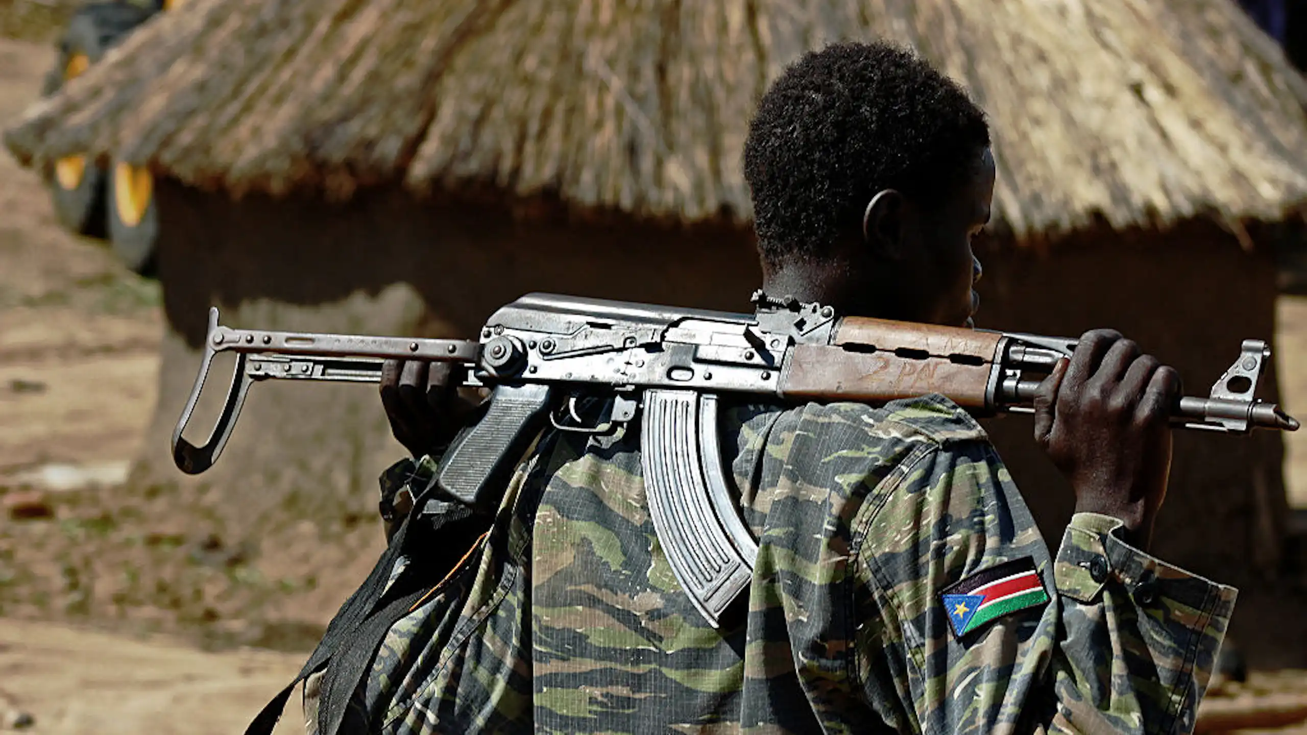 Man in camouflage uniform, holding a weapon, with a thatched mud hut in the background