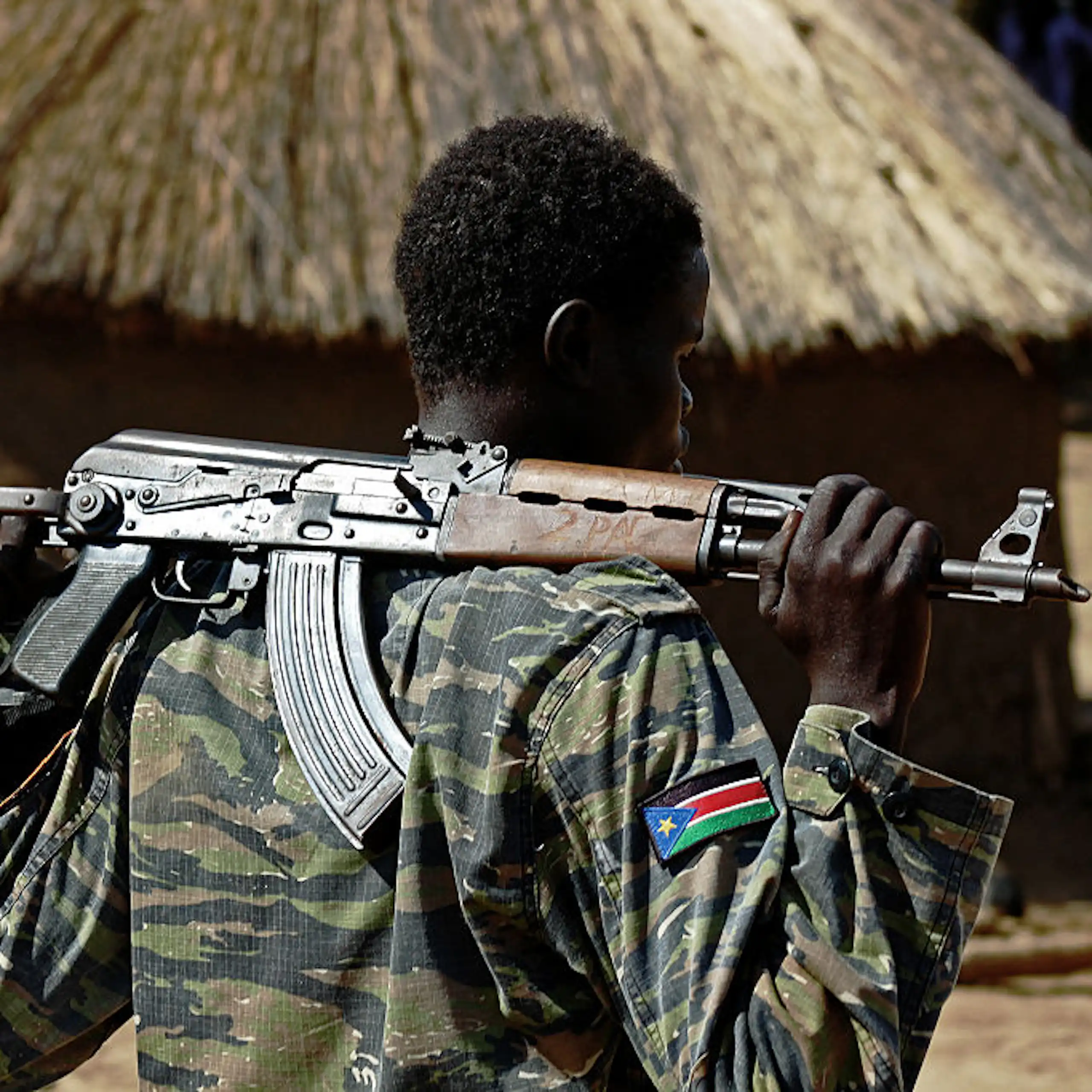 Man in camouflage uniform, holding a weapon, with a thatched mud hut in the background