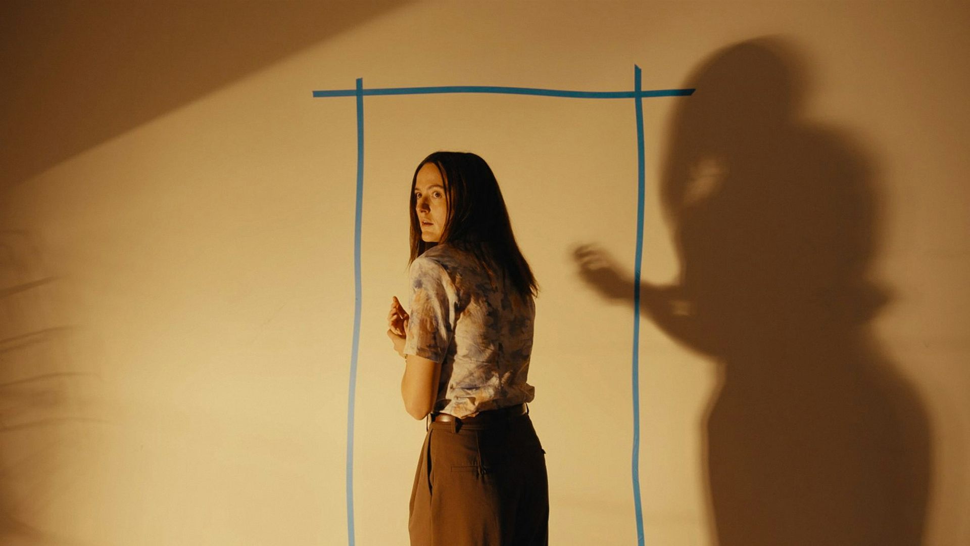 Woman looking at tape on a yellow wall in the shape of a door. A large shadow of her is cast behind.