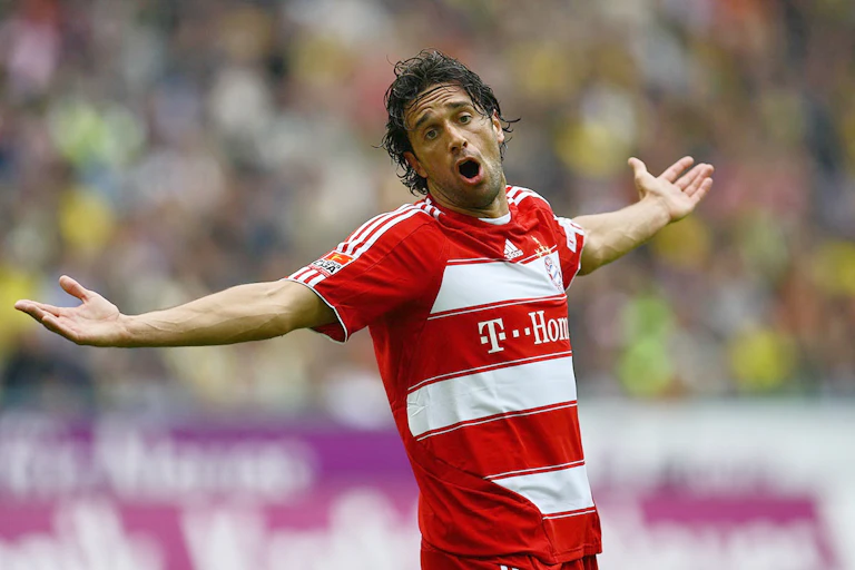 A close-up shot of a footballer in a red and white kit standing on a football pitch with outstretched arms.