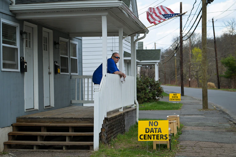 Man stands on front porch above yellow sign that says 'No Data Centers'