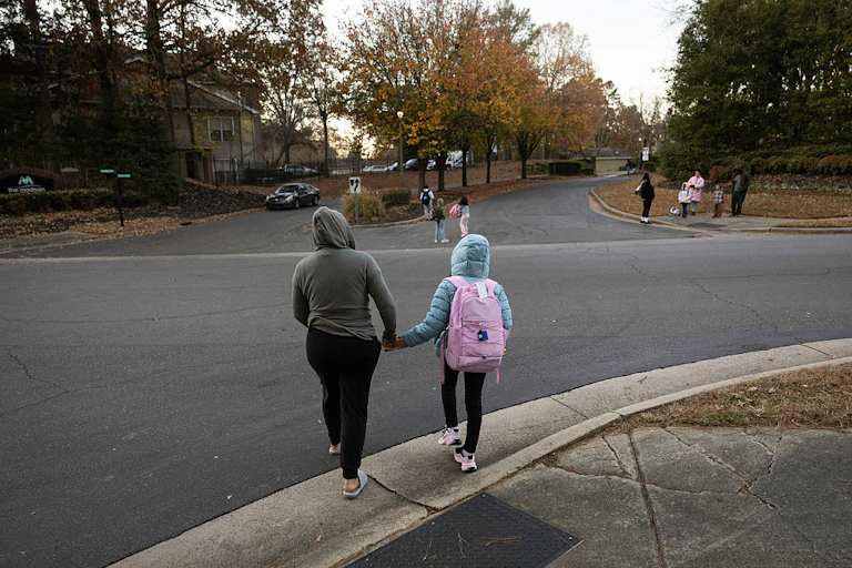 A person wearing a gray hoodie is seen from behind holding the hand of a child in a blue jacket and pink backpack as they walk on a street with trees that have orange and yellow and green leaves.