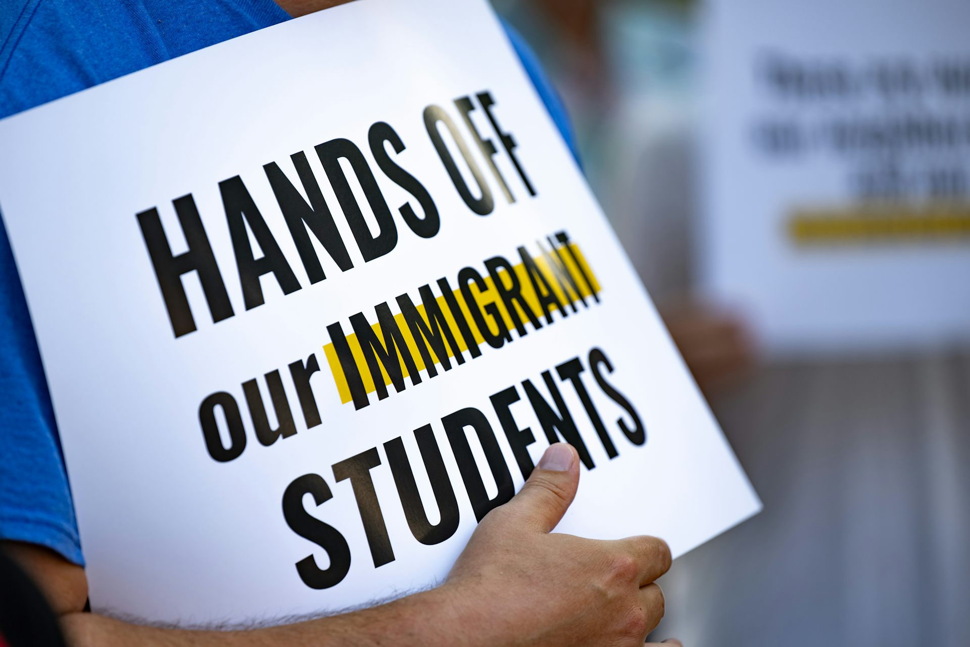 A white sign with black writing says 'Hands off our immigrant students'