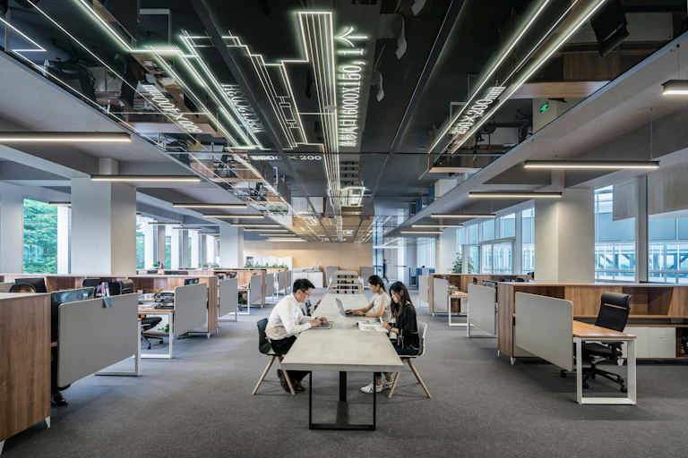Three workers with laptops sit at a long table in an otherwise empty office.