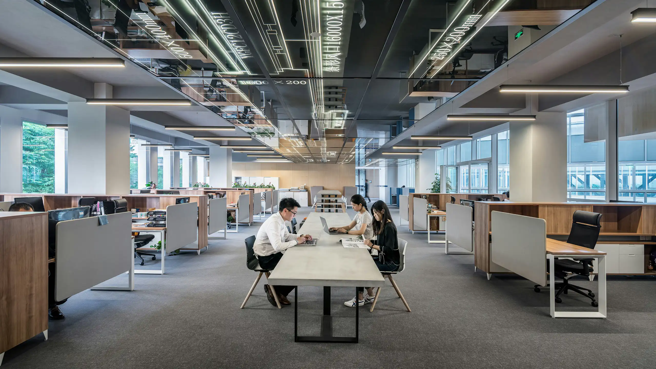Three workers with laptops sit at a long table in an otherwise empty office.