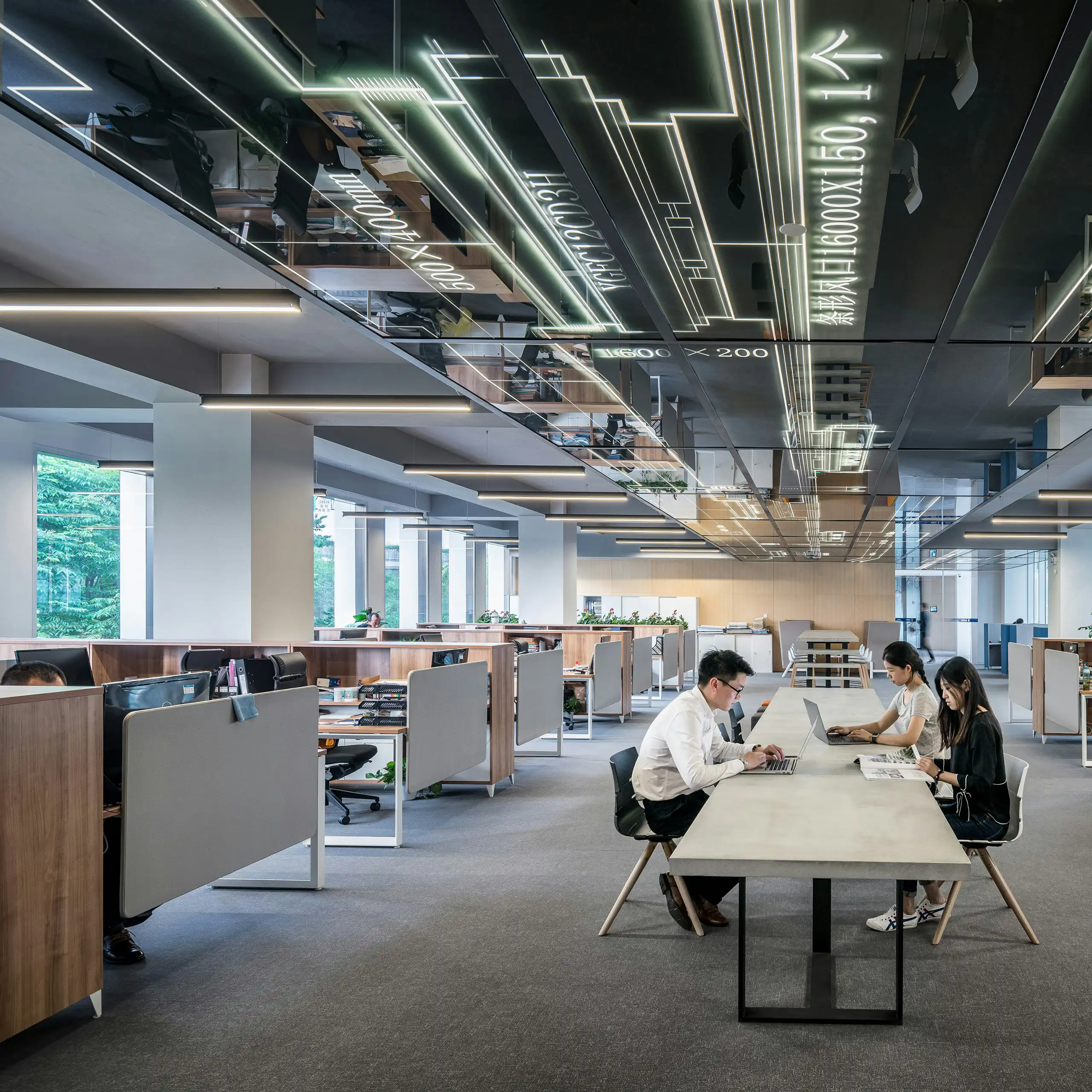 Three workers with laptops sit at a long table in an otherwise empty office.
