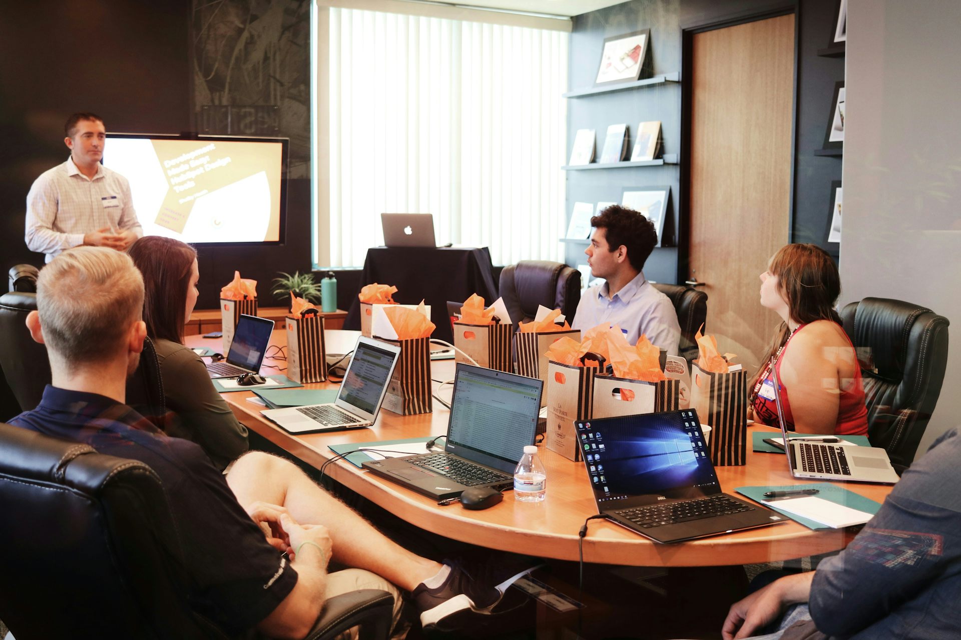 A group of workers sit around a table with laptops and gift bags on the table, listening to a presentation by a man in front of a screen.
