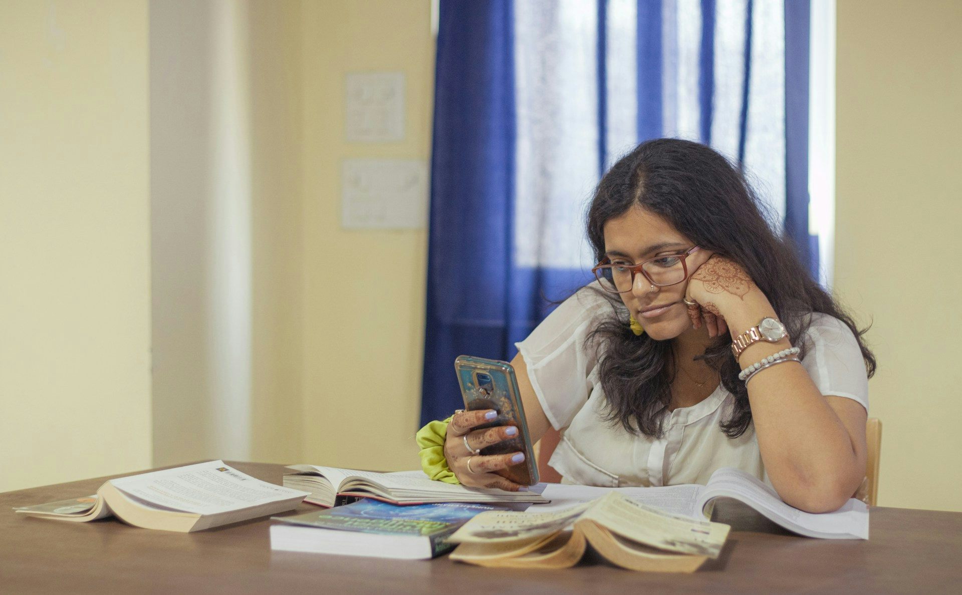 Una mujer con pelo largo y gafas mira fijamente su teléfono rodeada de libros sobre una mesa de estudio