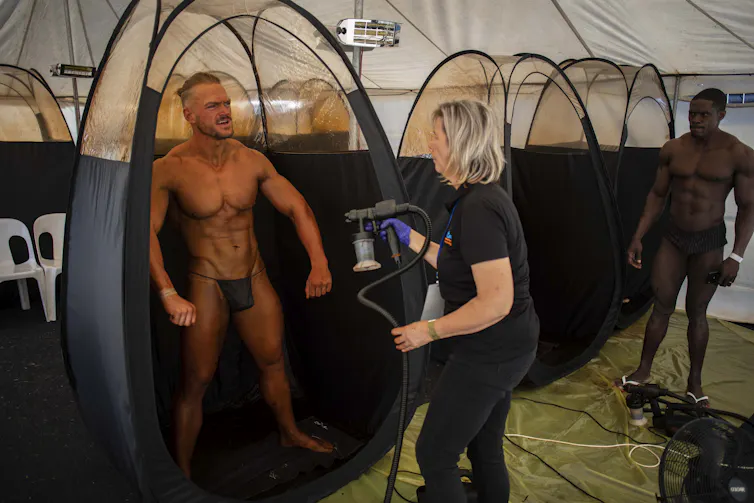 A man gets a spray on tan at a bodybuilding competition.