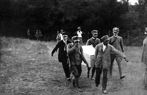 A group of uniformed men carry a coffin along a path out of woods