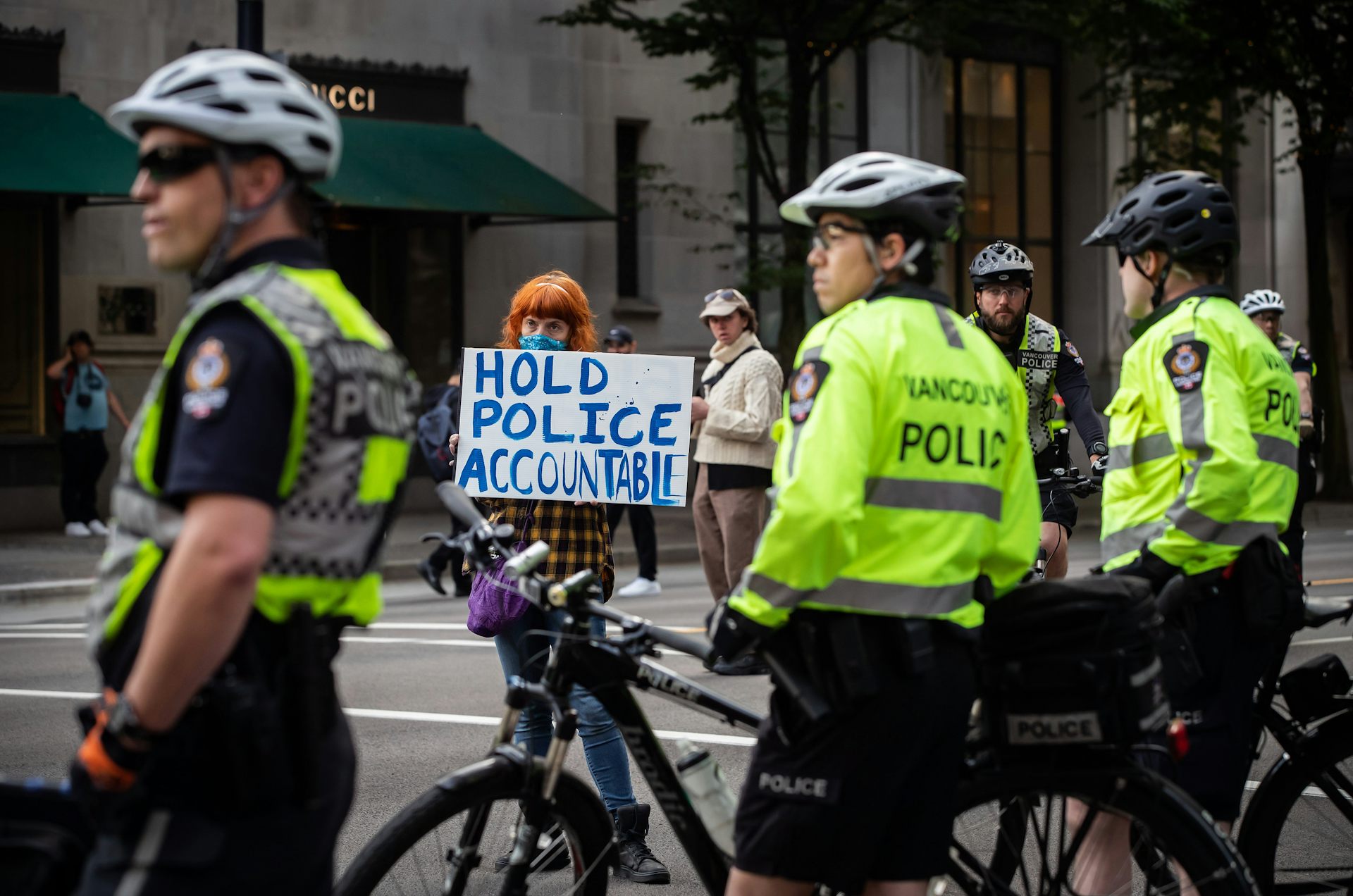 Three police officers are standing and holding their bicycles as they look on while a woman beyond them holds up a sign in protest.