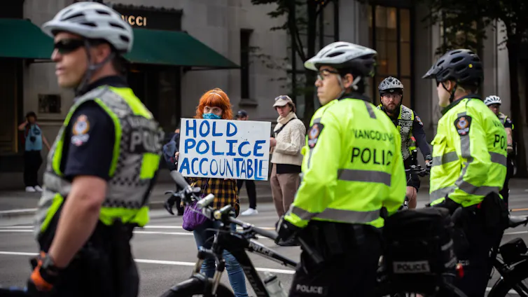 Three police officers are standing and holding their bicycles as they look on while a woman beyond them holds up a sign in protest.