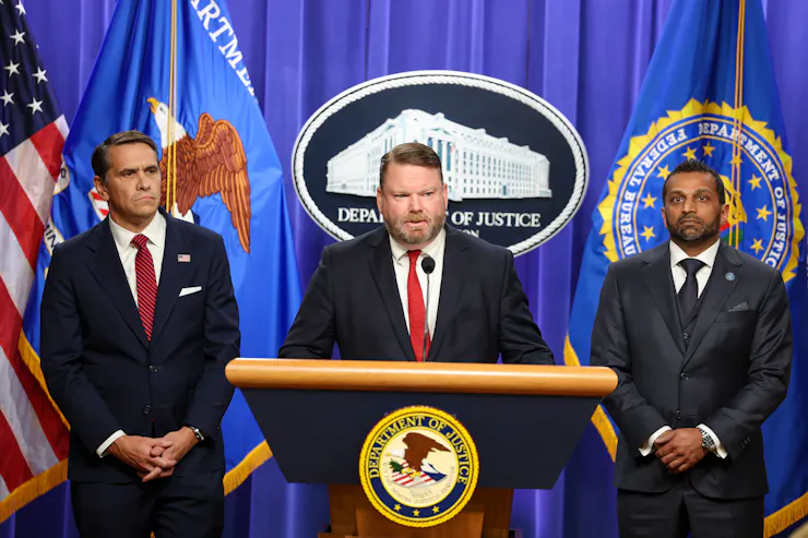 Three men standing in front of a curtain, flags and some official seals while one speaks at a lectern.