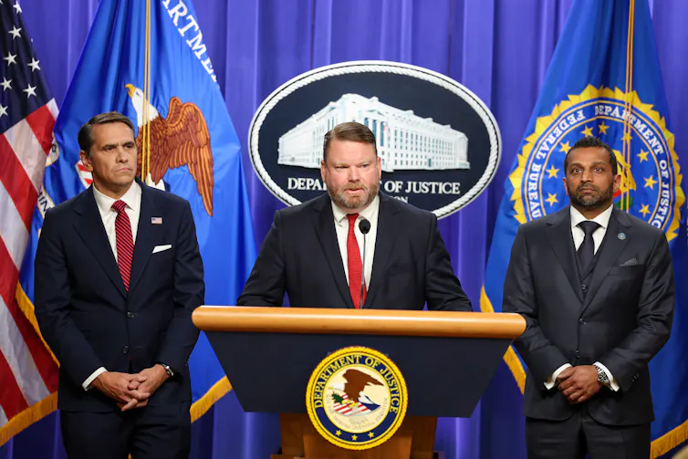 Three men standing in front of a curtain, flags and some official seals while one speaks at a lectern.