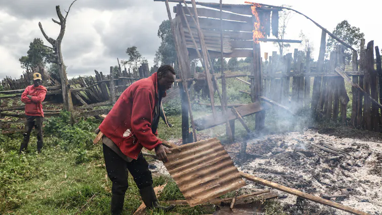 A man sifts through materials left after a building was destroyed, while part of the building burns in the background.