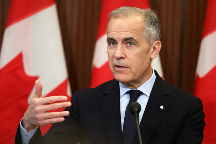 A man gestures as he speaks in front of a red and white flag.