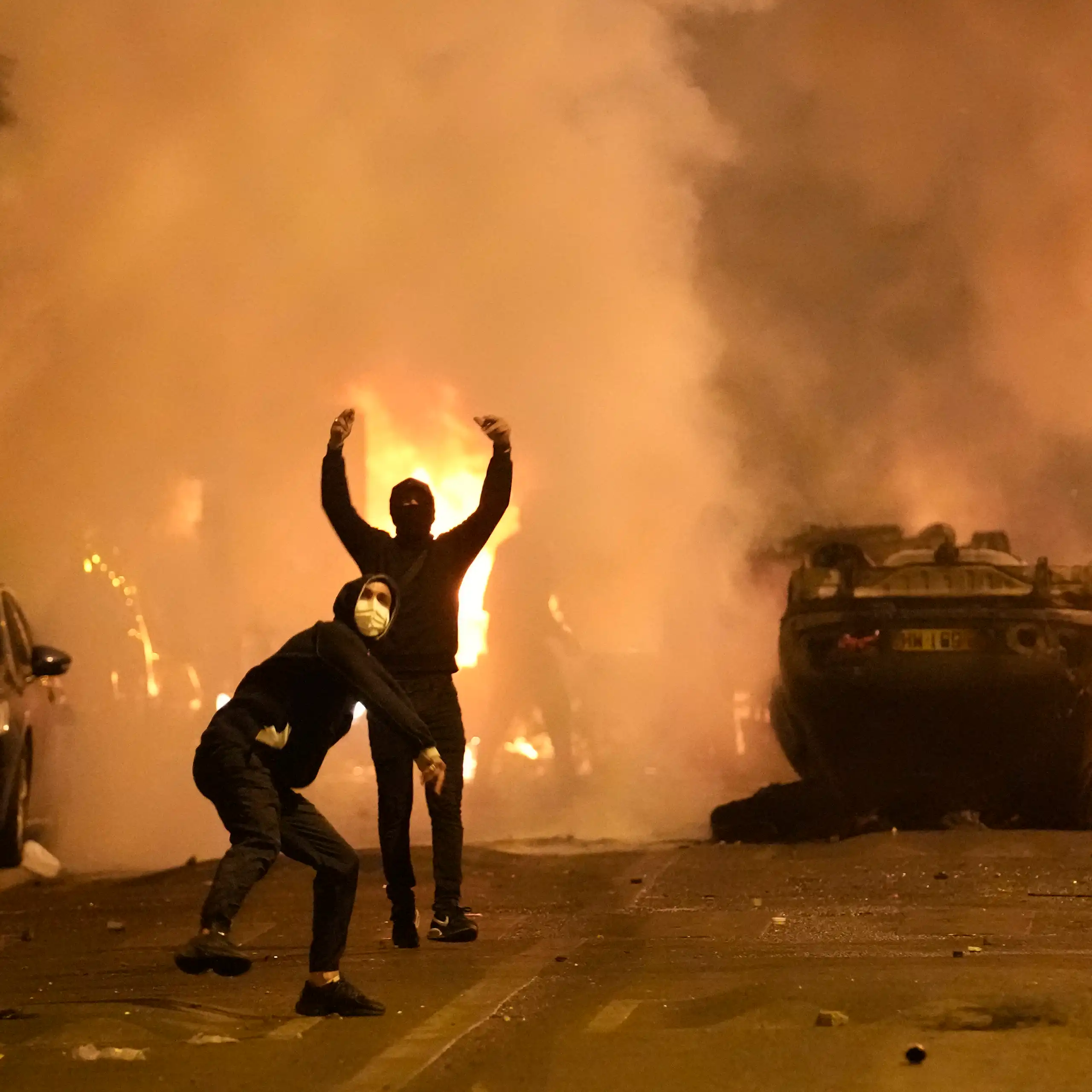 Des voitures renversées, dans un brasier, avec deux personnes à l'avant-plan, masquées
