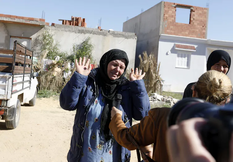 An elderly woman, wearing a headscarf, raises her hands in a gesture of helplessness in front of a group of people