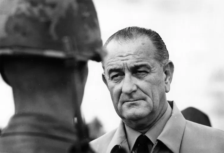 A balding man stands in front of a soldier who is wearing a helmet.