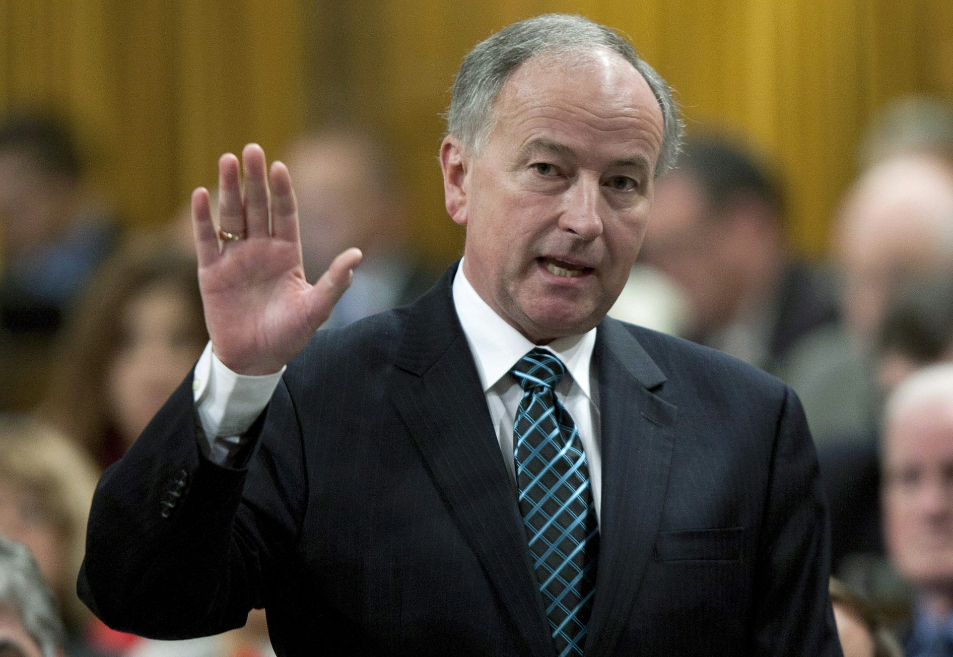 An older white man in a suit raises his hand while speaking