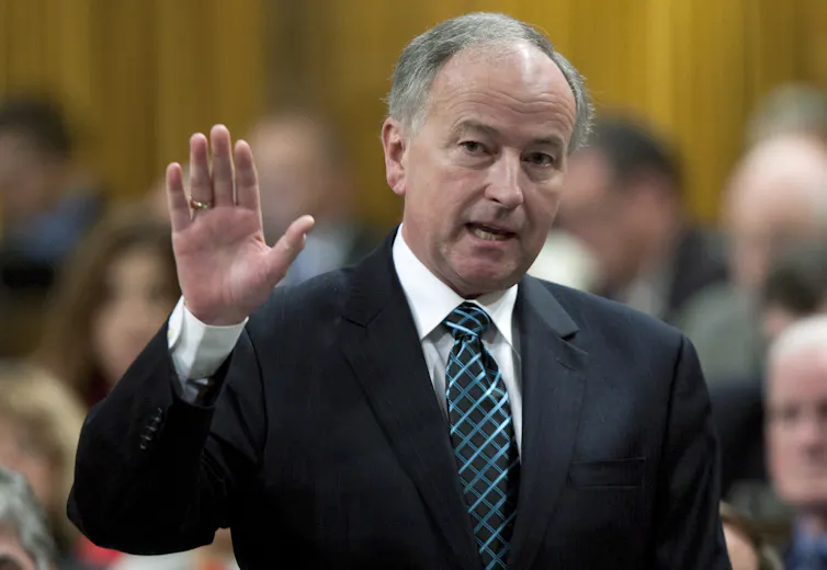 An older white man in a suit raises his hand while speaking