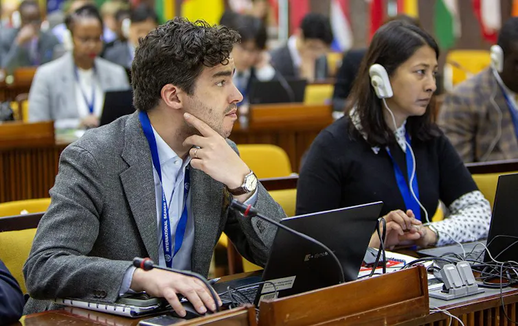 Image of two people sat at a desk looking upwards away from the camera. They have microphones in front of them and translation earpieces and there are more people in the background in similar positions.