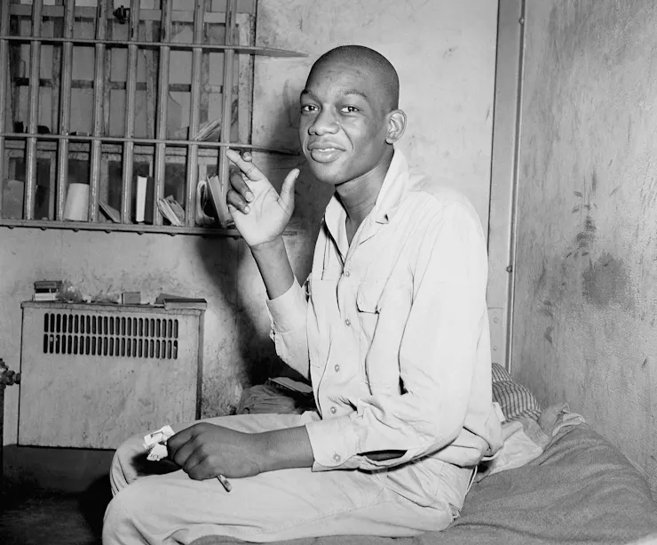 A young Black man sits in a prison cell and smiles.