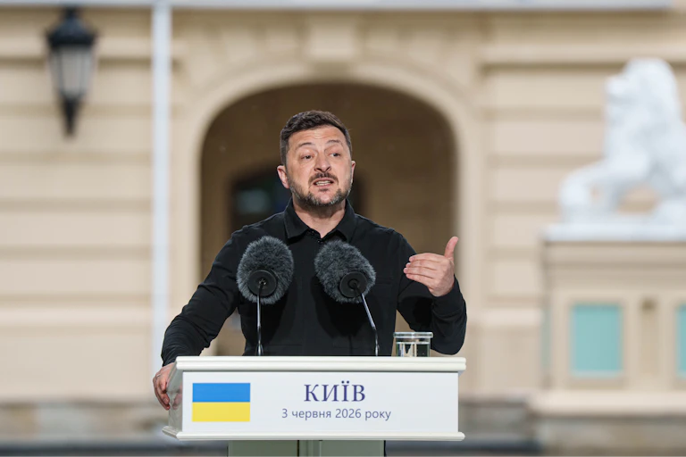 A middle-aged white man with a short beard in a black dress shirt speaks from behind a podium with the Ukrainian flag on it