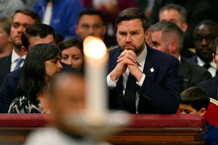 A brown-haired man looks ahead of him, hands clasped, as he leans his elbows on a padded railing.