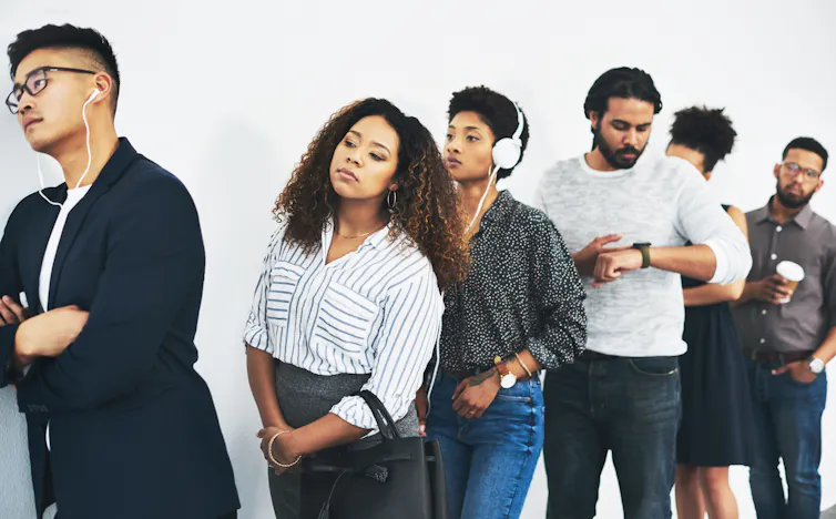 half dozen people standing in line against a wall, looking bored and impatient