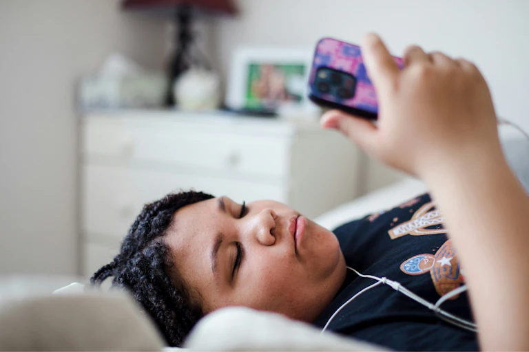 A teenager lying on their bed, looking at a phone and wearing headphones.