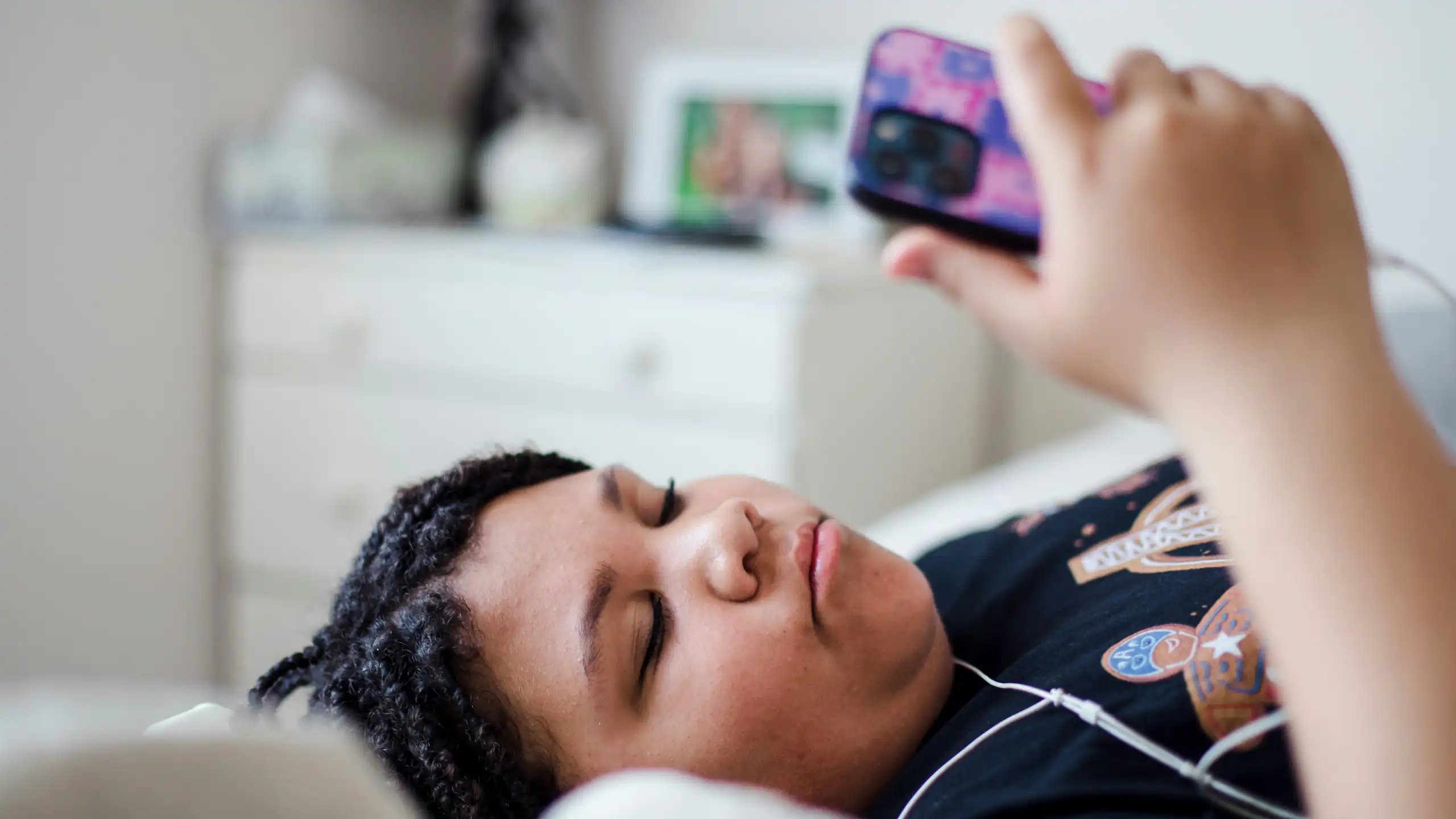 A teenager lying on their bed, looking at a phone and wearing headphones.