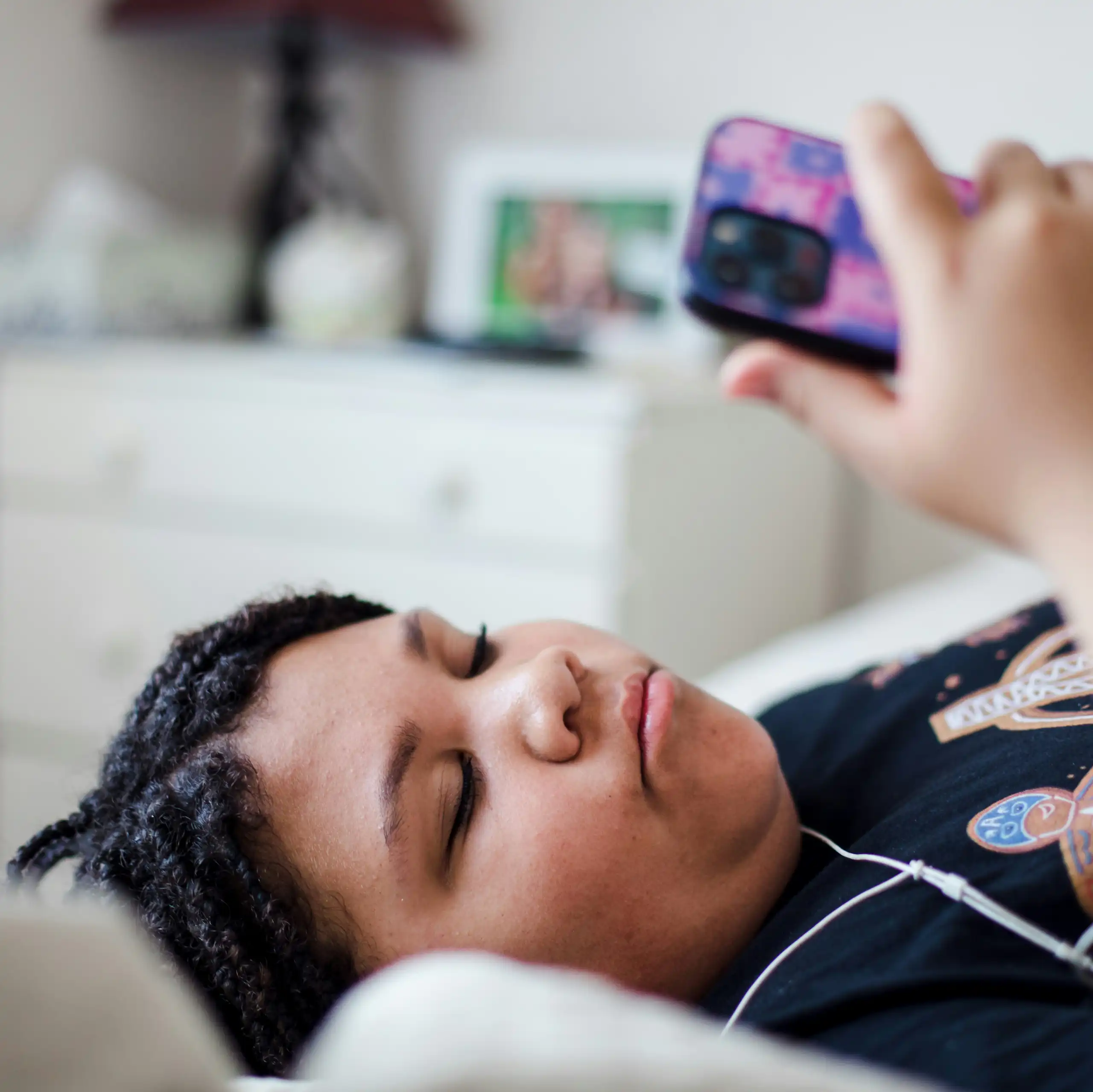 A teenager lying on their bed, looking at a phone and wearing headphones.
