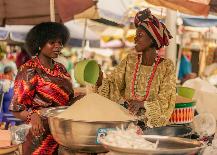 Two women over a bowl of dry rice at a market.