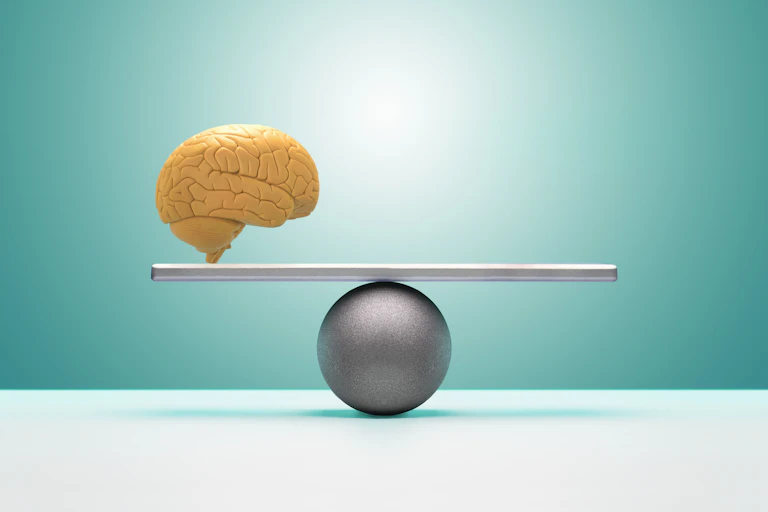 A yellow cartoon of a brain rests on one side of a perfectly balanced beam, set against a teal backdrop.