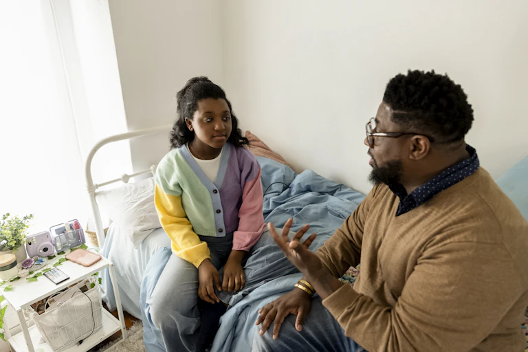 Father and adolescent daughter sitting on her bed, having a discussion.