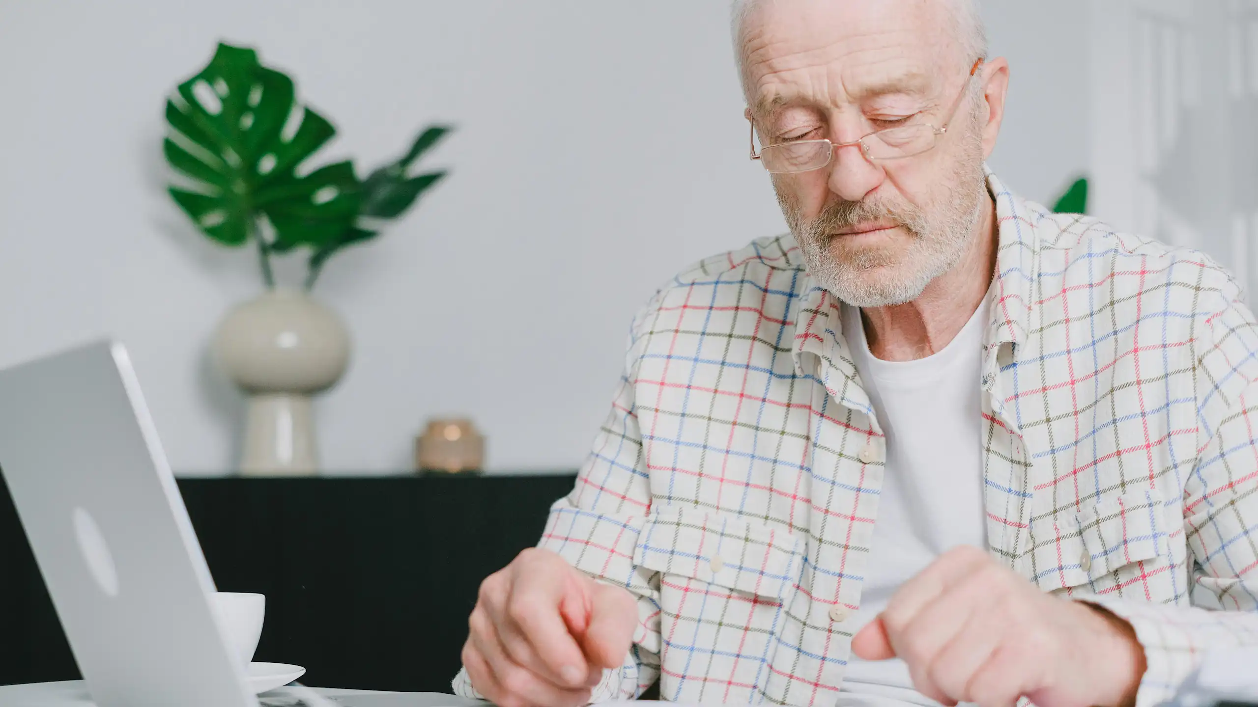 Elderly man with eyeglasses focused on paperwork at home, using laptop