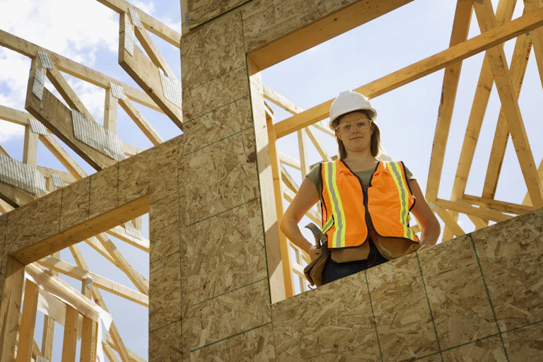 A young woman in hard hat and hi-vis vest stands on a building site.