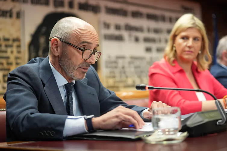 El empresario español Julio Martínez habla por un micrófono en una mesa mientras una mujer sentada cerca mira