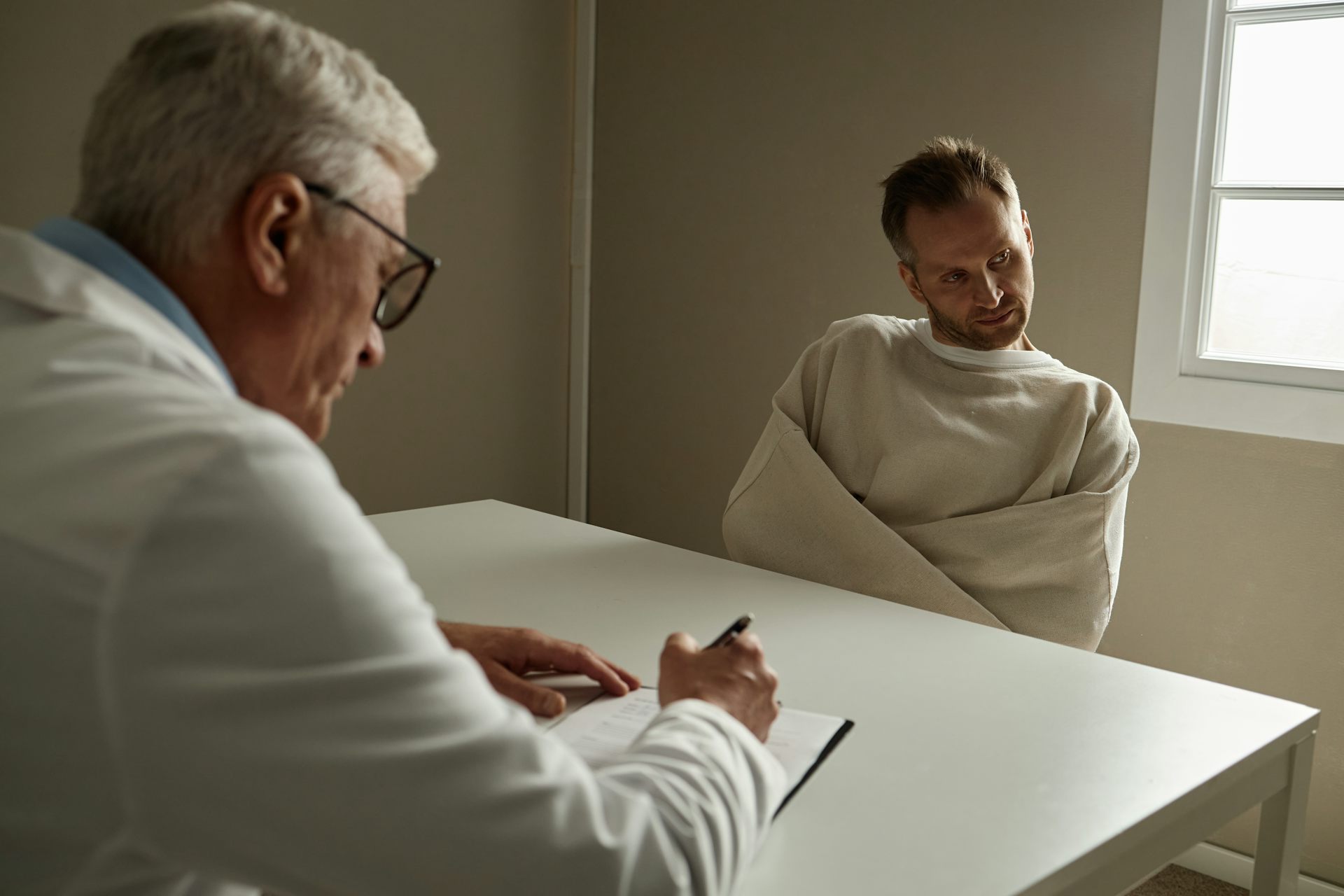 Un homme, l’air absent, est assis à une table, entravé par une camisole de force, face à un soignant plus âgé qui prend des notes.