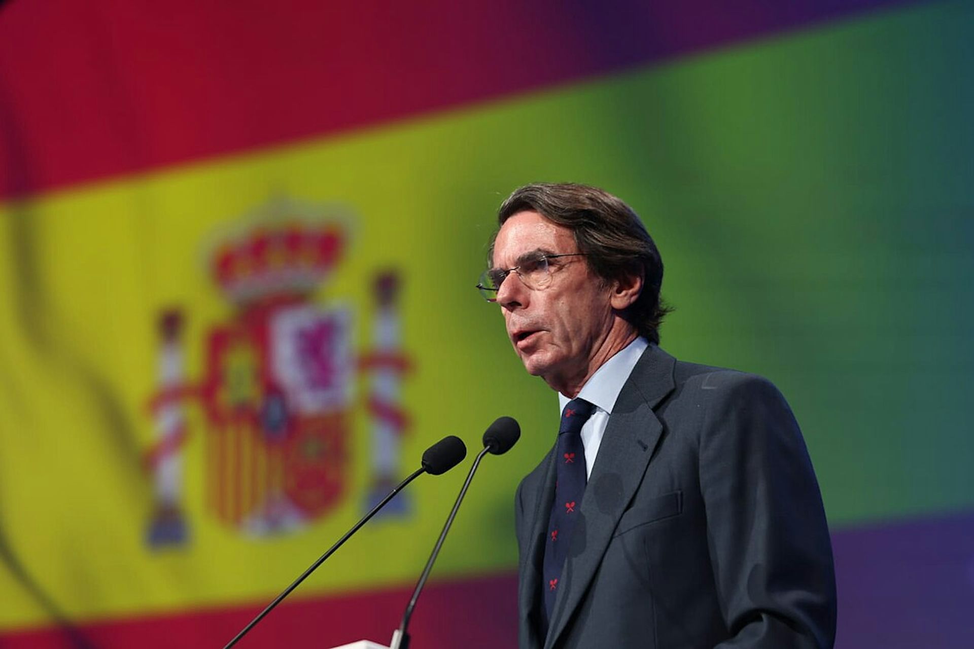 José María Aznar habla por el micrófono con la bandera de España de fondo.