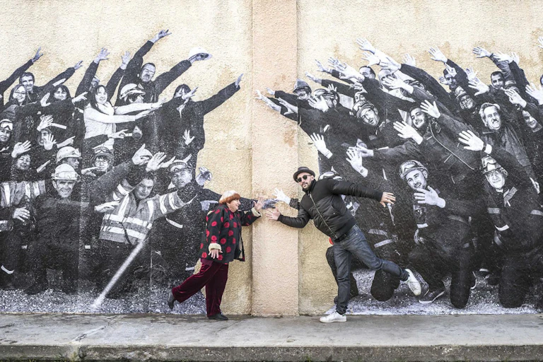 Un hombre y una mujer posan delante de un mural con obreros saludando.